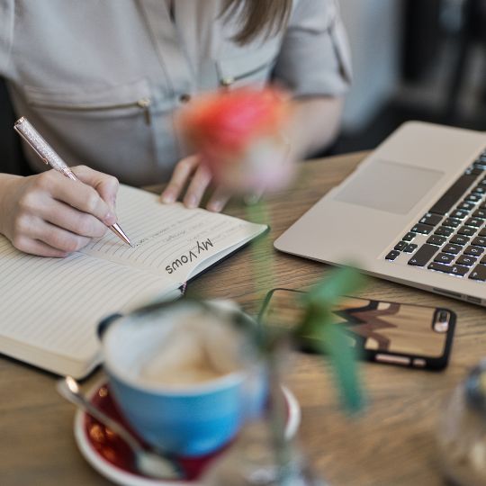 Woman using
a yellow pencil to writing vows in a notebook . A
blue cup of coffee on a red saucer, a laptop, a
mobile phone, and an out of focus pink rose in a
bud vase are also on the table