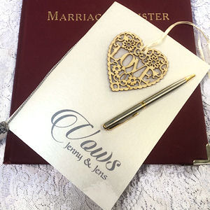 Cream card with the
words Vows Jenny & Jens on a burgundy marriage
register. Lying on the vows card is a wooden
filigree heart that includes the word love and a
silver pen lying on a pale carpet