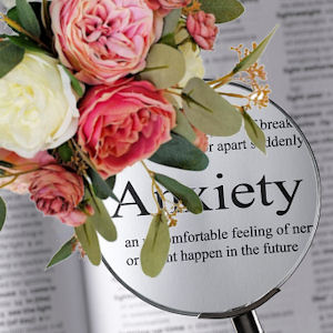 Bouquet of white and various shades of pink
peonies with gum leaves on a page with a magnifying
glass enlarging the word Anxiety
