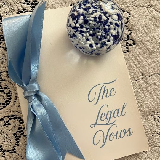 Vows folder
                      tied with pale blue ribbon. The words The Legal
                      Vows are on the front. The folder is on an ecru
                      lace tablecloth. A blue and white paperweight is
                      also on the table.