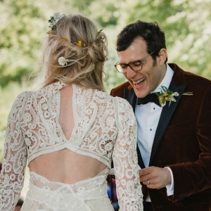 Groom laughing as he reads his vows. The
bride is seen only from the back. She is wearing a
lace dress with cut-outs.